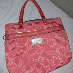 Röd axelväska från Coach - Säljer en snygg röd axelväska från Coach med silverfärgad logotypplatta och dragkedja framtill. Väskan har Coachs klassiska mönster och röda handtag. Perfekt för dig som vill ha en stilren och rymlig accessoar.