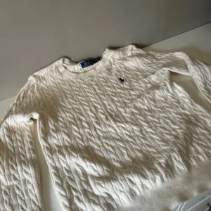 Vit kabelstickad tröja från Polo Ralph Lauren - Säljer en klassisk vit kabelstickad tröja från Polo Ralph Lauren. Tröjan har rund halsringning, lång ärm och den ikoniska broderade loggan på bröstet. Perfekt för lager-på-lager och stilrena outfits.