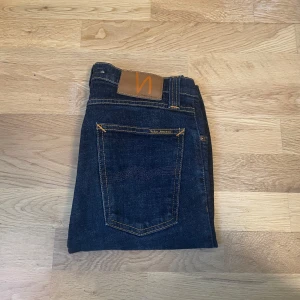 Mörkblå jeans från Nudie Jeans - Säljer ett par klassiska mörkblå jeans från Nudie Jeans med snygga kontrastsömmar och bakficka med diskret logga. Perfekta för dig som gillar stiliga jeans. Midjemått W28 och längd L34.