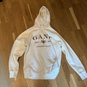 Vit hoodie från GANT - Säljer en vit hoodie från GANT med tryck framtill där det står 'GANT EST. 1949 CONNECTICUT' och en stjärndetalj. Tröjan har huva med dragsko och en stor magficka. Perfekt för en avslappnad stil. Har använts kanske 5 gånger