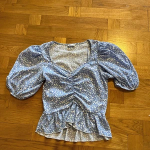 Blå blommig blus med puffärmar från Cubus - Supersöt blå blommig blus med v-ringning, puffiga trekvartsärmar och volang nertill. Blusen har rynkade detaljer framtill och är perfekt för vår och sommar. Lätt och luftig modell som ger en romantisk känsla.