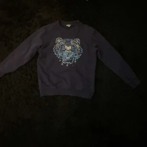 Kenzo Tröja  - Säljer en blå sweatshirt från Kenzo med det ikoniska tigerhuvudet broderat framtill i blå och vita nyanser. Tröjan har rund halsringning och långa ärmar. Perfekt för dig som gillar streetwear och vill sticka ut med en snygg logga.