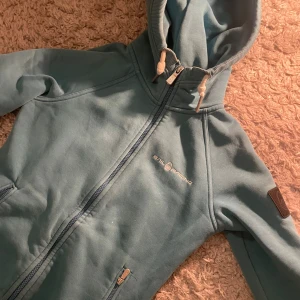 Ljusblå hoodie från Sail Racing - Ljusblå hoodie från Sail Racing med dragkedja och huva med vita snören. Broderad logga på bröstet och patch på ena ärmen. Mjuk och skön, perfekt för kyliga dagar.