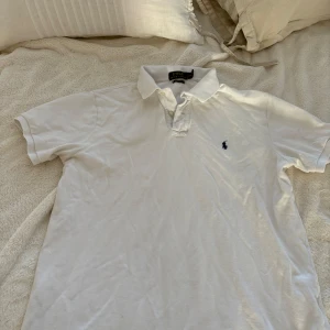 Vit pikétröja från Polo Ralph Lauren - Klassisk vit pikétröja från Polo Ralph Lauren med broderad logga på bröstet. Tröjan har korta ärmar, krage och knappar vid halsen. Custom fit-modell som passar perfekt till sommaren.