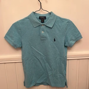 Ljusblå pikétröja från Polo Ralph Lauren - Säljer en ljusblå pikétröja från Polo Ralph Lauren med klassisk krage och två knappar framtill. Tröjan har korta ärmar och den ikoniska broderade loggan på bröstet. Perfekt för en stilren och avslappnad look.