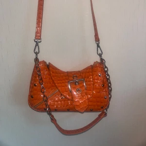 Orange axelväska med kedja och bälte - Säljer en cool orange axelväska med krokodilpräglat mönster och silverfärgade nitar. Väskan har en dekorativ bältesdetalj framtill, kedjehandtag och dragkedja på sidan. Perfekt för att lyfta vilken outfit som helst! Köpt på NA-KD för ett par år sedan, den är använd men i väldigt fint skick. Verkligen en av mina favoritväskor men använder den så sällan så tycker att det är dags för den att få komma till någon som använder den mer. 