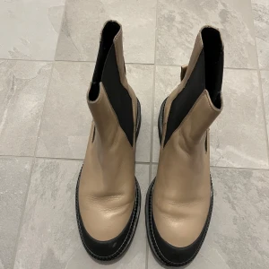 Beige och svarta boots med hög skaft - Säljer ett par beige boots med svarta detaljer och högre skaft. Skorna har rund tå, platt sula och elastiska svarta paneler på sidorna för enkel påtagning. Perfekta för dig som vill ha en stilren och modern look.