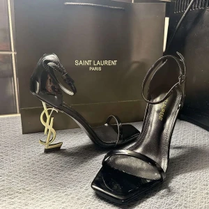Svarta Saint Laurent klackar med YSL-detalj - Säljer ett par svarta klackskor från Saint Laurent med ikonisk YSL-logga som klack. Skorna har öppen tå, smal rem över tårna och runt vristen. Tillverkade i glansigt skinn med fyrkantig tå och elegant design.