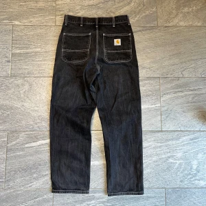 Svarta jeansbyxor från Carhartt - Säljer ett par svarta jeansbyxor från Carhartt med raka ben och stora fickor bak. Klassisk Carhartt-logga på ena bakfickan. Byxorna har normal passform och är tillverkade i slitstarkt denimtyg.