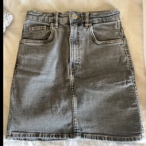 Grå jeanskjol - Säljer en klassisk grå jeanskjol med fem fickor och synliga sömmar. Kjolen har en rak passform, knapp och dragkedja framtill samt bälteshällor. Perfekt att styla med en enkel topp eller hoodie.