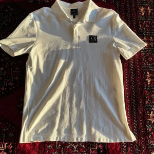 Vit pikétröja från Armani Exchange - Säljer en stilren vit pikétröja från Armani Exchange med klassisk krage och korta ärmar. Tröjan har en svart AX-logga på bröstet och är tillverkad i bomull. Aldrig använd! 