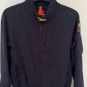 Mörkblå vindjacka från Parajumpers - Snygg mörkblå vindjacka från Parajumpers med hög krage och dragkedja framtill. Jackan har en patch på ärmen och praktiska fickor med dragkedja. Perfekt för blåsiga dagar och enkel att matcha med olika outfits.