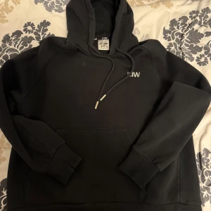 Svart hoodie från ICANIWILL - Säljer en svart hoodie från ICANIWILL med justerbar huva och ficka framtill. Tröjan har långa ärmar och en liten vit logga på bröstet. Perfekt för en avslappnad och sportig stil.