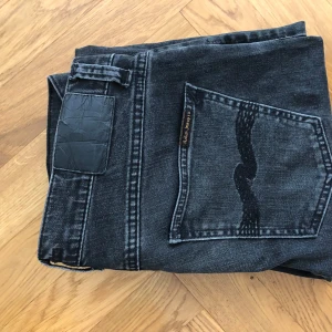 Svarta jeansbyxor från Nudie Jeans - Säljer ett par svart/gråa jeansbyxor från Nudie Jeans med klassisk femficksdesign och svarta sömmar. Jeansen har normal passform och raka ben. Perfekta för dig som gillar stilrena och enkla jeans. Storlek: 30/30