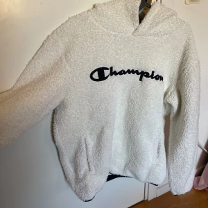Vit teddy-hoodie från Champion - Supermjuk och fluffig vit hoodie från Champion med broderad logga framtill. Tröjan har huva och är tillverkad i teddy-material som håller dig varm och bekväm. Perfekt för kyliga dagar och en avslappnad stil.