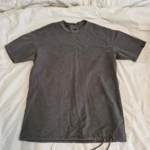 Grå t-shirt från Gymshark med snörning Stl S - Säljer en grå t-shirt från Gymshark med korta ärmar och rund hals. T-shirten har en unik snörning nertill och diskret logga på ärmen. Perfekt för dig som gillar stilrena träningskläder.