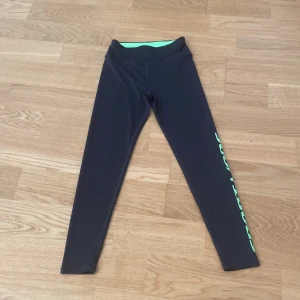 Svarta sportleggings från H&M med grön text - Svarta sportleggings från H&M med hög midja och grön insida i midjan. Ena benet har tryckt texten 'SPORT MODE' i neon-grönt längs sidan. Perfekta för träning och har en tight passform.