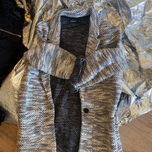 Svartvit kappa med knytband - Säljer en svartvit mönstrad kappa med bred krage och knytband i midjan. Jackan har en klassisk kavajkrage och är långärmad. Perfekt för dig som vill ha en stilren och trendig look.