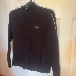 Svart boss jacka med dragkedja  - Säljer en svart jacka från BOSS med hög krage och hel dragkedja framtill. Jackan har diskret BOSS-logga på bröstet och vita detaljer på ärmarna. Perfekt för en stilren och sportig look. Säljs eftersom jag har bytt stil. Ny pris runt 1800 kr