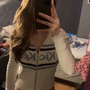 Vit ribbad ziptröja från Kickers - Säljer en vit ribbad ziptröja från Kickers med blå broderad logga och ränder över bröstet. Tröjan har lång ärm och är tillverkad i ett stickat material som ger en snygg och bekväm passform. Perfekt för dig som gillar sportig och retro stil. Den är i storlek uk 8