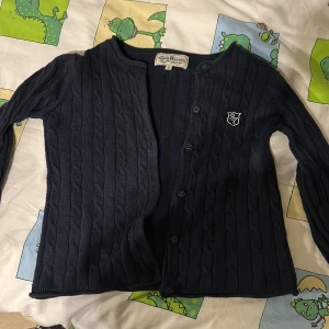 Mörkblå kabelstickad kofta från Hampton Republic - Mörkblå kabelstickad kofta från Hampton Republic med knappar framtill och broderad logga på bröstet. Koftan har lång ärm och en klassisk, något croppad passform. 