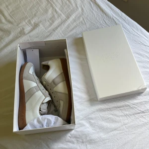 Maison margiela - Säljer ett par stilrena vita sneakers med beige mockadetaljer och klassisk brun gummisula. Skorna har snörning och en retroinspirerad design som passar perfekt till många outfits. Levereras i originalkartong.