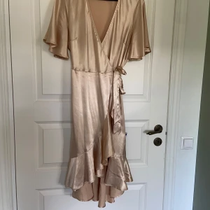 Champagnefärgad omlottklänning från Pieces - Säljer en elegant champagnefärgad omlottklänning från Pieces i glansigt satinmaterial. Klänningen har korta vida ärmar, volangdetaljer nertill och knyt i midjan. Perfekt för speciella tillfällen.