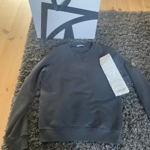 Mörkgrå sweatshirt från Stone Island Junior i storlek 160cm - Säljer en stilren mörkgrå sweatshirt från Stone Island Junior. Tröjan har rund halsringning, långa ärmar och klassisk logotyp-patch på vänster ärm. Perfekt för en avslappnad och trendig look. Tröjan kostade 1949 nypris och jag köpte den på nk i Göteborg. 9/10 skick