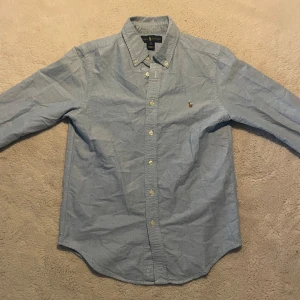 Ljusblå skjorta från Ralph Lauren - Klassisk ljusblå skjorta från Ralph Lauren med broderad logga på bröstet. Skjortan har lång ärm, knappar framtill och button-down krage. Perfekt för en stilren look. Storlek 14-16 men passar dig ifrån 155-168 