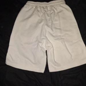 Vita shorts med svart hjärta och A-logga - Säljer ett par vita shorts från AMI Paris med elastisk midja och snörning. På vänster ben finns en stor svart hjärta-A-logga. Shortsen är raka i modellen och har en mjuk bomullskänsla, perfekta för sommaren.