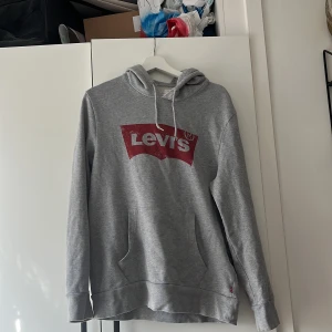 Grå hoodie från Levi's - Säljer en klassisk grå hoodie från Levi's med röd logga framtill. Tröjan har huva med dragsko och en stor magficka. Perfekt för en avslappnad stil.