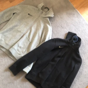 Nike hoodie i svart och grått - Två snygga Nike hoodies i storlek L, en i svart och en i ljusgrå. Båda har dragkedja, huva och fickor framtill. Perfekta för träning eller chill, med klassisk Nike-logga på bröstet.