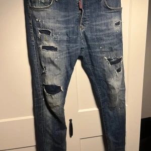 Dsquared2 slim her jeans med slitningar - Säljer ett par blå Dsquared2 Skinny Dan jeans i storlek 48 med tydliga slitningar och hål på framsidan. Jeansen har klassisk femficksdesign, smal passform och detaljer som färgstänk och patch bak.