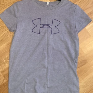 Grå t-shirt från Under Armour - Säljer en grå t-shirt från Under Armour med deras klassiska logga tryckt på bröstet. T-shirten har korta ärmar och är i en loose fit-modell, perfekt för träning eller vardag. Materialet är mjukt och bekvämt bomull.