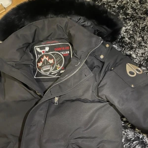 Svart dunjacka med pälskant från Moose Knuckles - Säljer en svart dunjacka från Moose Knuckles med pälskant på huvan och klassisk logga på ärmen. Jackan har dragkedja, knappar och flera fickor. Perfekt för kalla dagar och riktigt snygg design.