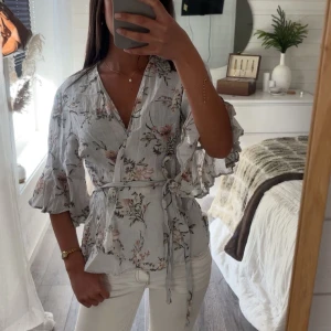 Blommig omlottblus med volangärm - Säljer en ljusblå omlottblus med blommigt mönster och volangärmar. Blusen har knytband i midjan och v-ringning🌊☀️🤍🫧
