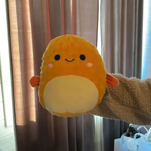 Gul Squishmallows plyschleksak - Supermjuk och kramvänlig gul Squishmallows plyschleksak med orange fenor och vitt ansikte. Den har ett sött leende och runda rosa kinder. Perfekt som gosedjur eller dekoration i rummet.