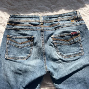 Blå jeansbyxor med slitningar - Ett par klassiska blå jeansbyxor med fem fickor och orangea sömmar. Byxorna har låg midja och flare ben. Perfekta för en casual stil.🥰