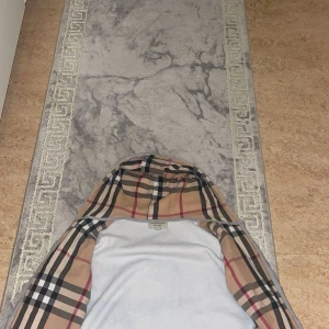 Grå hoodie med Burberry-inspirerad rutig huva - Grå zip-hoodie med klassisk rutig huva i beige, svart och rött. Jackan har dragkedja framtill och snörning i huvan. Perfekt för en avslappnad och stilren look.