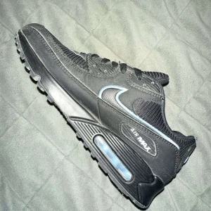 Svarta Nike Air Max sneakers med blå detaljer - Säljer ett par svarta Nike Air Max sneakers med ljusblå detaljer runt swoosh-loggan och Air Max-texten. Skorna har en synlig luftbubbla i sulan och är tillverkade i en mix av syntet och mesh. Perfekta för dig som gillar klassisk streetstil.