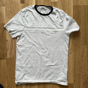 Ljusgrå t-shirt från Tommy Hilfiger med broderad logga - Säljer en ljusgrå t-shirt från Tommy Hilfiger med broderad logga över bröstet och en vit rand. T-shirten har svart ribbad krage och klassisk passform. Perfekt för en stilren och enkel look.