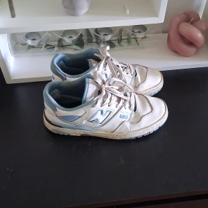 New Balance 550 sneakers i vitt och ljusblått - Säljer ett par New Balance 550 sneakers i vitt med ljusblå detaljer. Skorna har snörning, platt sula och rund tå. Perfekta för dig som gillar klassisk streetwear-stil och vill ha bekväma sneakers till vardags.