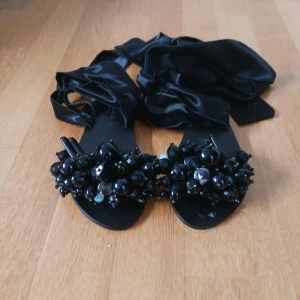 Svarta sandaler med pärldetaljer från Elina Linardaki - Unika svarta sandaler från Elina Linardaki med stora svarta pärlor och stenar framtill. Skorna har breda band i satin som knyts runt vristen för en snygg look. Perfekta för dig som vill sticka ut med accessoarer. Strlk 38 helt nya aldrig använda. Handgjorda och kostar runt 2500kr nya. Pris går att diskuteras.