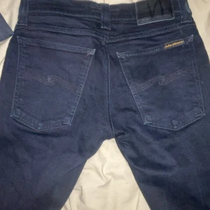 Svarta jeans från Nudie Jeans - Säljer ett par svarta jeans från Nudie Jeans i dammodell, storlek 25/30. Jeansen är tillverkade i denim och har klassisk femficksdesign med Nudie-logga på bakfickan och svart läderpatch i midjan. Kontakta för minsta lilla. Dom är helt nya 