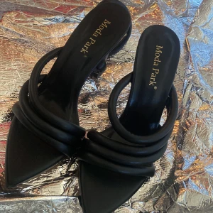 Svarta klackskor från Moda Park - Säljer ett par svarta klackskor från Moda Park med öppen häl och spetsig tå. Skorna har flera rundade remmar över foten och en elegant design som passar till många tillfällen.