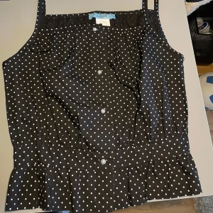 Brun prickig linnetopp med volang - Söt brun linnetopp från Gina Tricot med vita prickar, knappar framtill och volang nertill. Smala axelband och lätt rynkad upptill för en feminin look. Perfekt för varma dagar!