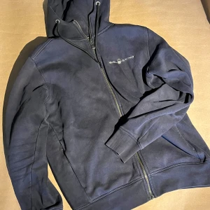 Mörkblå hoodie från Sail Racing - Mörkblå hoodie från Sail Racing med dragkedja och huva. Jackan har broderad logga på bröstet och är fodrad med mjuk fleece på insidan. Perfekt för kyliga dagar och enkel att matcha med andra plagg.