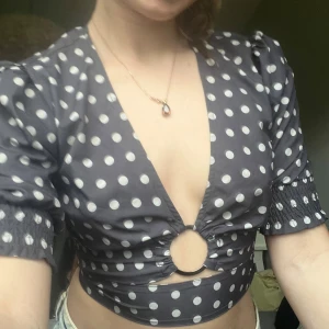 Svart prickig croppad blus från NA-KD x AFJ - Säljer en svart croppad blus med vita prickar från NA-KD x AFJ. Toppen har djup v-ringning, korta puffärmar och en snygg ringdetalj framtill. Perfekt för dig som gillar trendiga och unika toppar!