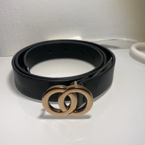 Svart bälte med guldfärgat spänne från Gina Tricot - Stilrent svart bälte från Gina Tricot med ett snyggt guldfärgat spänne i form av två ringar. Perfekt accessoar för att lyfta din outfit. Bältet är gjort i skinnimitation och har flera hål för justerbar passform.