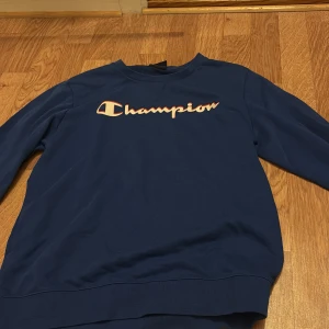 Blå sweatshirt från Champion - Säljer en blå sweatshirt från Champion med logga i vitt och rött på bröstet. Tröjan har rund hals och långa ärmar, perfekt för en avslappnad stil. Passar både tjejer och killar som gillar streetwear.